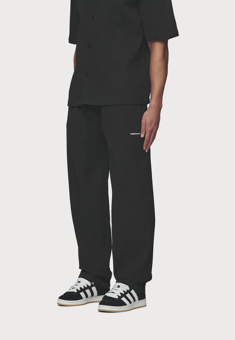 Black, ribbed short-sleeve shirt with buttons and loose-fitting pants. Shoes are black suede with white stripes and laces.