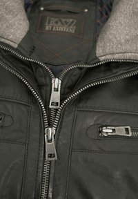 Schwarze Lederjacke mit grauem Stoffkragen, ausgestattet mit mehreren silbernen Reißverschlüssen und gestepptem Innenfutter. Auffällige Nähte im Inneren.