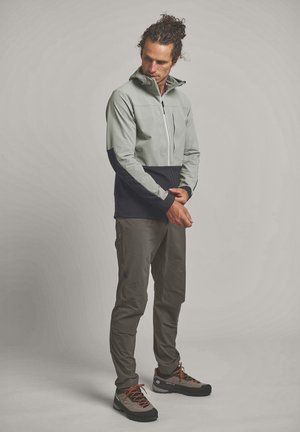 Mann justiert den Ärmel einer zweifarbigen Jacke, trägt graue Hose und braune Wanderschuhe, steht vor hellem einfarbigem Hintergrund.