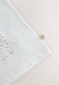 Fehér pamutszövet sima textúrával, arany vonal mintázattal és egy kis bézs címkével, amelyen az "Little Bird" felirat olvasható, a szélén rögzítve.