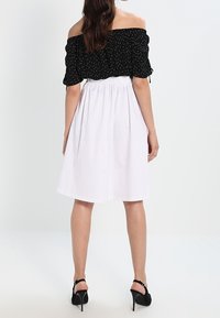 Top nero off-the-shoulder con pois bianchi, abbinato a una gonna bianca a vita elastica di lunghezza al ginocchio. Sono indossate scarpe nere con tacco alto.