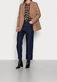 Blazer marrone su misura sopra una blusa con stampa leopardata, abbinato a jeans a gamba larga blu scuro e stivali alla caviglia neri con tacco.