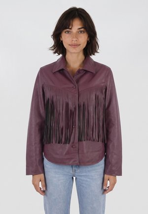 Donna che indossa una giacca di pelle viola con lunghe frange sul petto e jeans azzurri, in piedi davanti a uno sfondo chiaro e uniforme.