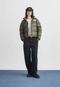 Νεαρό άτομο που φοράει ελιά πράσινο μπουφάν puffer, μαύρο παντελόνι, μαύρα παπούτσια και μαύρο καπέλο, στέκεται με τα χέρια στις τσέπες πάνω σε μπλε πάτωμα.