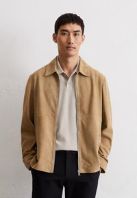 Tan suède jack met een relaxte kraag, ritssluiting en zijzakken. Gecombineerd met een lichtgrijze textuur polo en donkere broek.