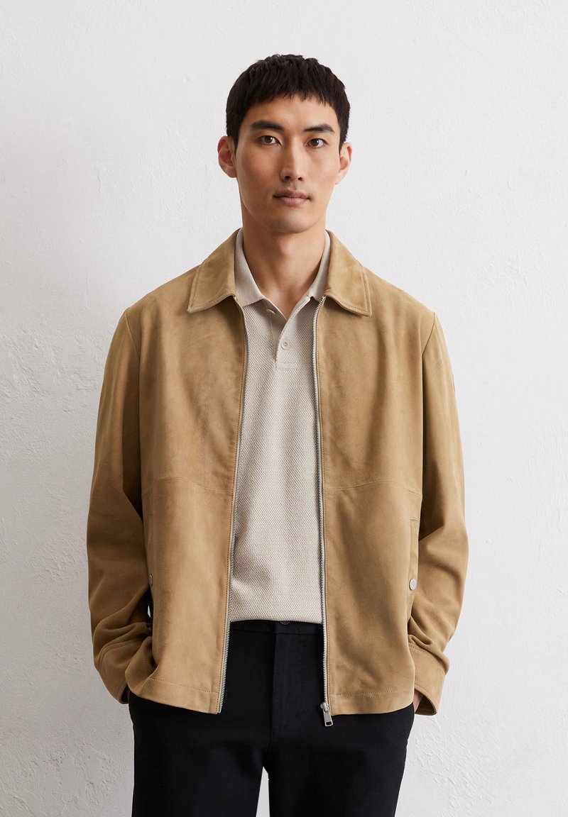 Tan suède jack met een relaxte kraag, ritssluiting en zijzakken. Gecombineerd met een lichtgrijze textuur polo en donkere broek.