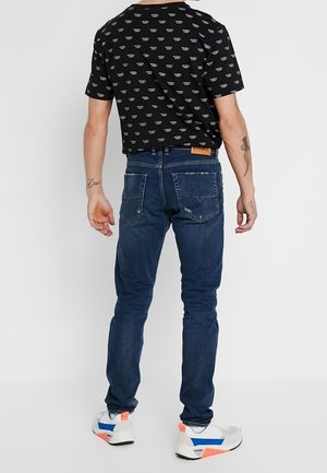 Mann, der ein schwarzes gemustertes T-Shirt, blaue Slim-Fit-Jeans mit Gesäßtaschen und weiße Sneaker mit blauen und orangefarbenen Akzenten trägt, steht mit dem Rücken zur Kamera.