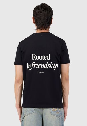 Man draagt een zwart T-shirt met de tekst "Rooted by friendship" op de rug, gecombineerd met lichtblauwe spijkerbroek, staand met de rug naar voren.