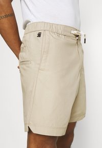 Man wearing beige drawstring shorts with side and back pockets, a small black logo on the waist, and a white t-shirt.