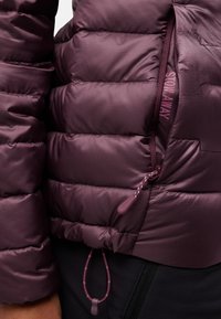 Giacca puffa viola con sezioni trapuntate, dotata di tasca con zip e orlo regolabile. Tessuto liscio e lucido con una texture delicata.