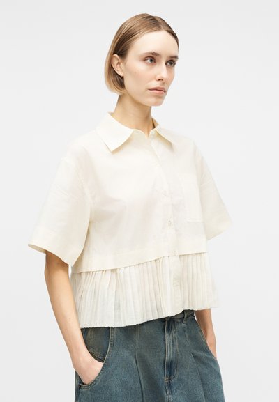 Γυναίκα με κοντά ξανθά μαλλιά που φοράει μια εκρού κοντομάνικη μπλούζα με κουμπιά και πιέτες στο τελείωμα και μπλε τζιν, με τα χέρια στις τσέπες, ουδέτερο φόντο.