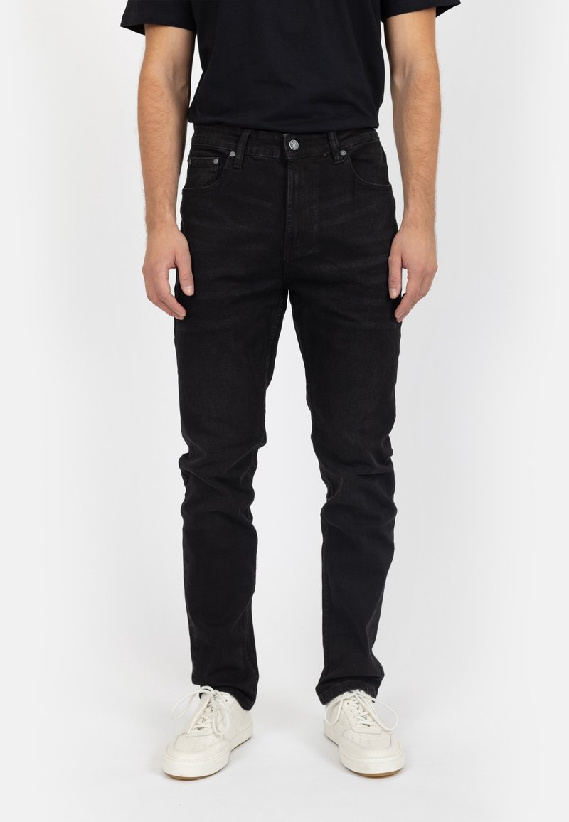 Homme portant un jean noir slim et des baskets blanches, debout devant un fond blanc uni.