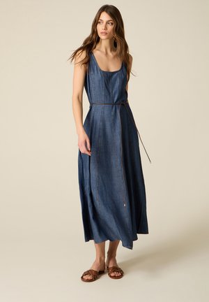 Frau trägt ärmelloses blaues Jeans-Maxikleid mit dünnem braunem Gürtel und braunen flachen Sandalen, steht vor beigem Hintergrund.