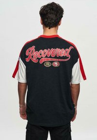 Sort-hvid t-shirt med korte ærmer og røde accenter, med teksten "Recovered" i fed skrift på ryggen og et logo fra San Francisco 49ers.