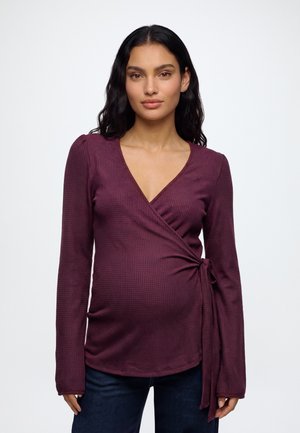 Femme enceinte aux cheveux ondulés foncés portant un haut cache-cœur à manches longues violet profond noué à la taille et un jean bleu foncé, debout sur un fond blanc.