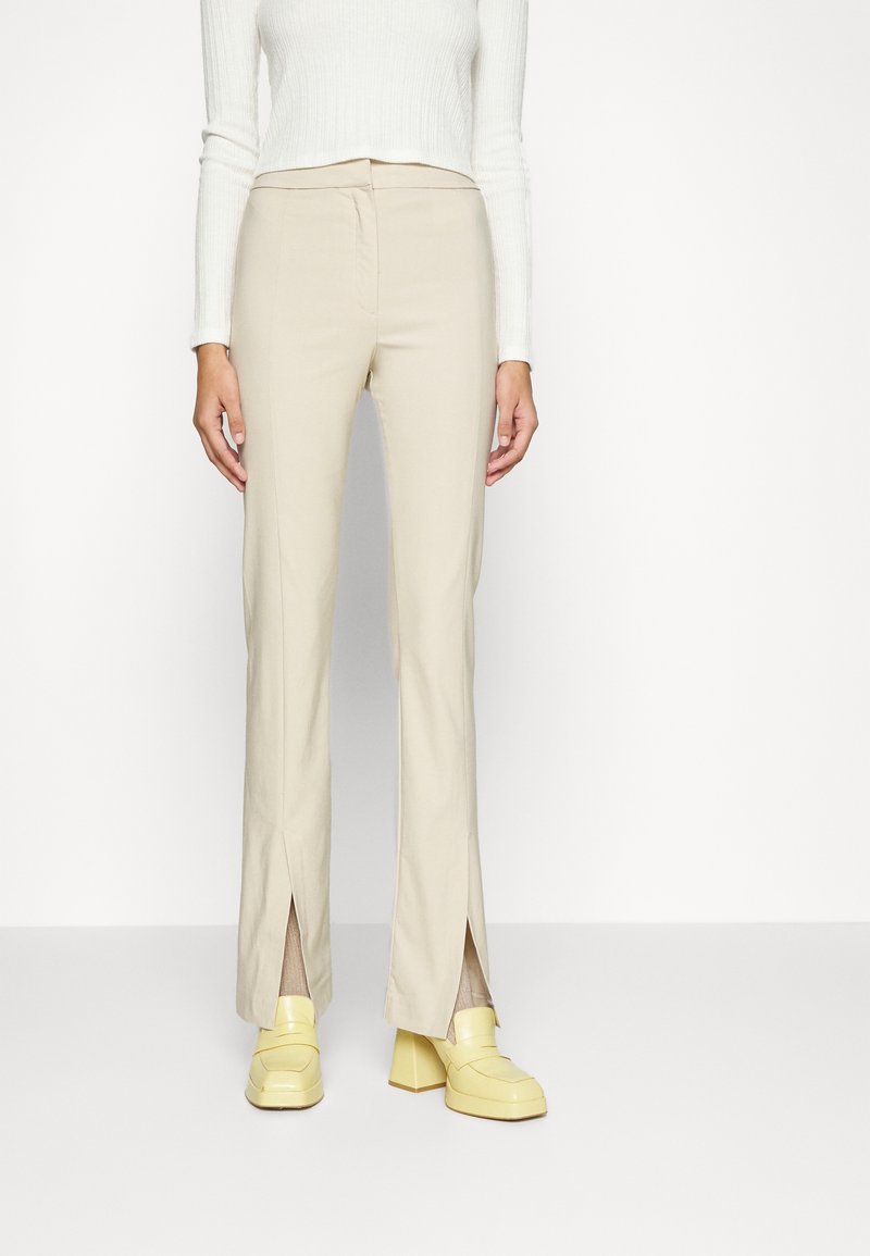 Femme portant un pantalon beige taille haute avec des fentes à l'avant des jambes, associé à des chaussures à talons épais jaunes et un haut blanc à manches longues côtelé.
