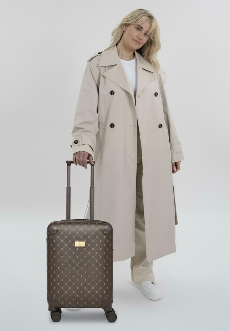 Valise marron à motifs avec des accents dorés, coque rigide, poignée rétractable et roues lisses, assortie à un trench-coat clair et des baskets blanches.