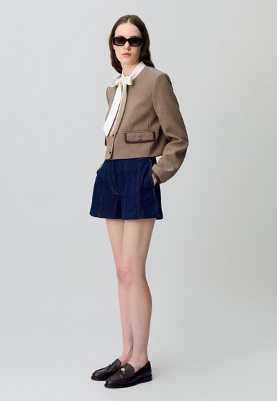 Blazer court à carreaux marron avec un nœud papillon blanc, associé à un short en denim foncé et des mocassins noirs. Le mannequin porte des lunettes de soleil.