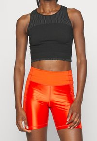 Czarny sportowy crop top z teksturowaną siateczką, w połączeniu z intensywnie pomarańczowymi dopasowanymi szortami z błyszczącą powierzchnią i detalami szwów.