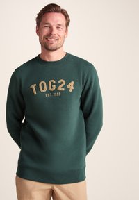 Green sweatshirt featuring textured gold lettering "TOG24 EST. 1958," with a crew neckline and ribbed cuffs and hem. Smooth cotton fabric.