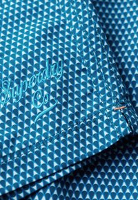 Blauwe stoffen met patroon heeft een geometrisch ontwerp met kleine driehoeken. Geborduurd "Superdry Co" logo in lichtblauw, met oranje stikdetails.