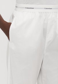 Weiße Calvin Klein-Hose mit einem breiten Bund mit Logo, seitlichen Taschen und einem glatten, leichten Stoff. Sauberes Design mit minimalen Details.