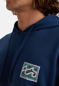 Sudadera de color azul marino hecha de un tejido suave con un bolsillo frontal. Presenta un diseño bordado cuadrado en tonos verde azulado, marrón y blanco. Cordonera ajustable.