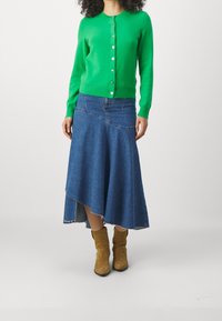 Zelený pletený cardigan s kulatým výstřihem a zapínáním na knoflíky, v kombinaci s asymetrickou džínsovou sukní a béžovými kotníkovými botami.