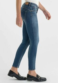 Jean skinny en denim bleu foncé avec cinq poches, surpiqûres jaunes et patch en cuir au dos. Associés à des mocassins noirs à talons bas.