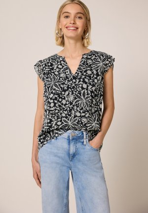 Hymyilevä nainen, jolla on hopeiset renkaanmuotoiset korvakorut, mustavalkoinen kukallinen hihaton toppi ja vaaleansiniset farkut, poseeraa yhdellä kädellä taskussa.