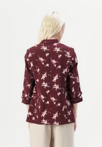 Blusa floreale bordeaux con maniche a tre quarti, caratterizzata da piccoli fiori rosa e bianchi, colletto con scollatura a V e dettaglio della cucitura posteriore.