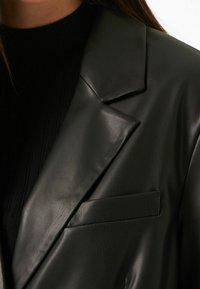 Svart läderblazer med ett skärpkrage, slät textur och en enstaka bröstficka. Tyget har en subtil lyster.