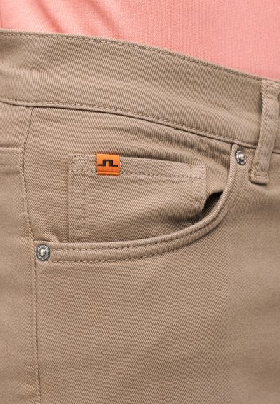 Τσέπη μπεζ παντελονιού με πορτοκαλί ετικέτα λογότυπου, ασημένιες τρούκας, λεπτομερής ραφή, φορεμένη με ροζ υφασμάτινο τοπ ορατό πάνω από τη ζώνη.
