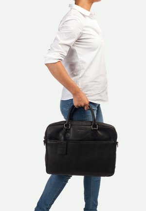 Sacoche d'ordinateur portable en cuir noir avec deux poignées supérieures, texture lisse, poche avant et étiquette de bagage. Elle a une forme rectangulaire et un logo discret.