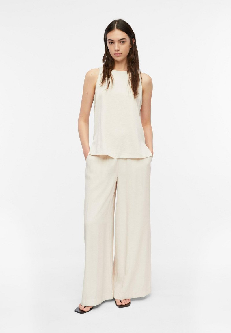 Young woman wearing sleeveless cream top and wide-leg cream pants with hands in pockets, standing on white background.