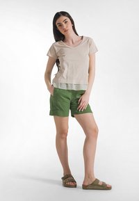 Camiseta de cuello en V color beige claro superpuesta sobre una blusa verde menta, combinada con pantalones cortos verdes. Modelo con sandalias planas marrones. Textura suave.