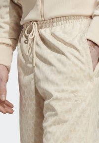 Béžové jogger kalhoty se jemným vzorem listů, elastickým pasem se stahovací šňůrkou a vyšitým logem na boku.