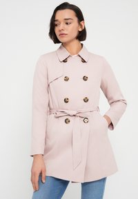 Ροζ παλτό τύπου τραντς με διπλή σειρά κουμπιών, ζώνη στη μέση που δένει, και γιακά με εγκοπή. Το ύφασμα φαίνεται λείο με ματ φινίρισμα.