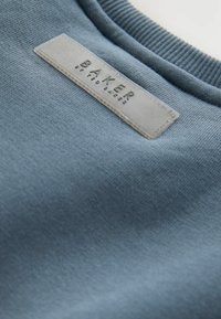 Blaues Sweatshirt mit weichem Stoff. Verfügbar mit einem gerippten Halsausschnitt und einem rechteckigen Stoffetikett, auf dem "BAKER" in hellgrau geprägt ist.
