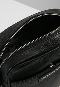 Černá kožená crossbody taška s texturovaným povrchem, vybavená stříbrným zipem, hladkým vnitřkem a značkovou nálepkou na přední straně.