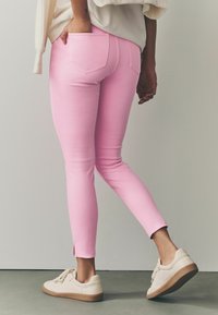 Jeans ajustados de color rosa con corte slim, que cuentan con bolsillos traseros y aberturas en los tobillos, combinados con zapatillas crema sobre una superficie lisa.