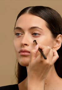 Makeup application with a hand holding a small brush, applying product under the eye of a person with fair skin and long dark hair, neutral background.