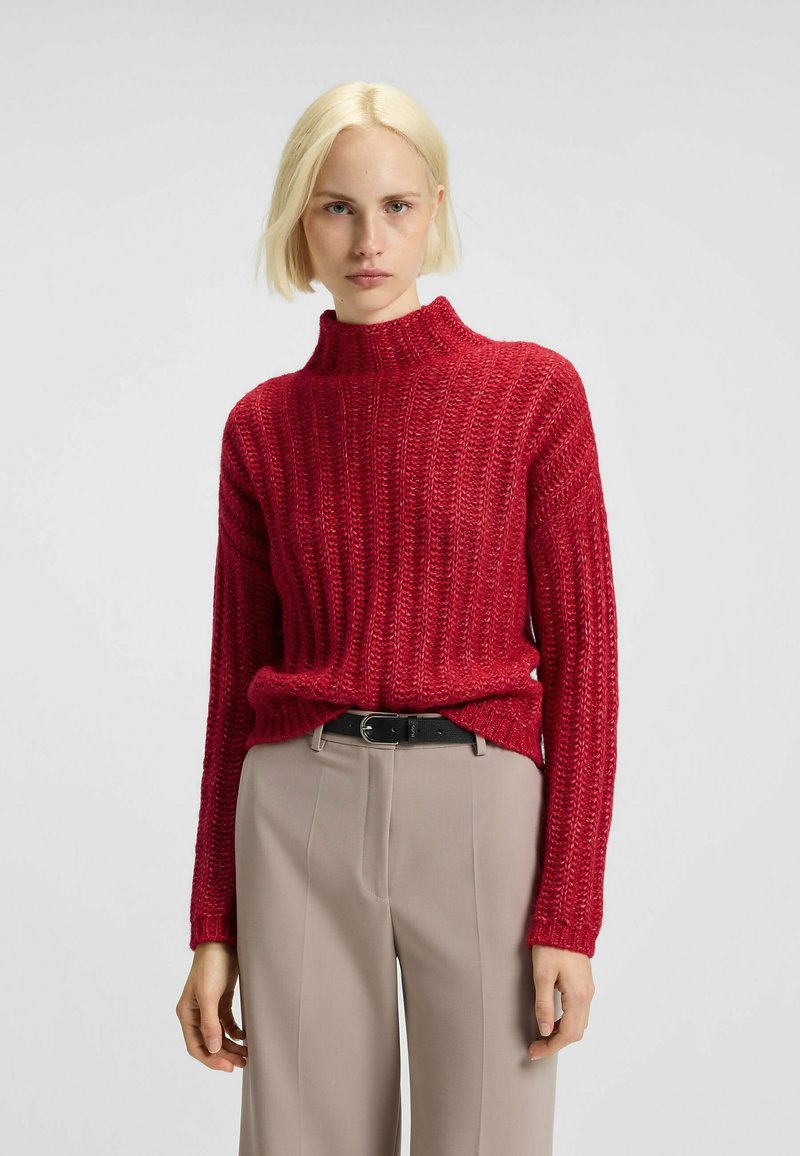 Rode geribbelde trui met een hoge halslijn, met textuurstof. Gecombineerd met beige broek en een zwarte riem.