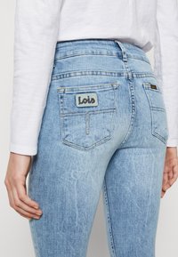 Vaaleansiniset farkut, joissa on korkea vyötärö ja takataskut. Vasemmassa taskussa on harmaa "Lois" -merkki, ja farkuissa on hieno haalistuminen ja kulutuksia.