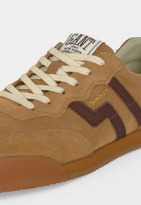 Basket en daim marron clair avec lacets blancs, bandes latérales en zigzag marron foncé et une étiquette en tissu portant la mention GANT sur la languette.