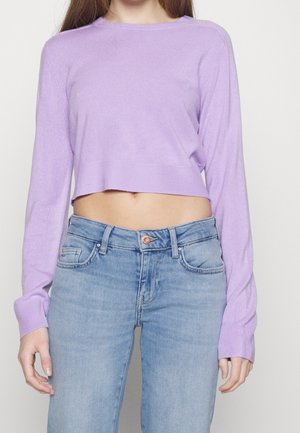 Personne portant un pull court à manches longues lavande associé à un jean taille haute bleu clair sur un fond uni.