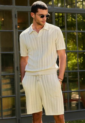 Mand iført cremefarvet strikket kortærmet skjorte med krave og matchende shorts, stående med hænderne i lommerne foran glasdøre.