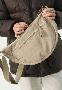 Beige Umhängetasche aus weichem, strukturiertem Stoff. Besitzt eine geschwungene Form, einen Reißverschluss und einen verstellbaren Tragegurt. Zwei Vordertaschen.