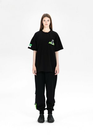 Joven mujer de pie, con una camiseta negra de gran tamaño con gráfico y pantalones negros a juego con detalles verdes, zapatos negros texturizados.