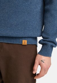 Blå ribbad stickad tröja med strukturerad yta och en Timberland-etikett vid fållen, kombinerad med bruna byxor i tyg.